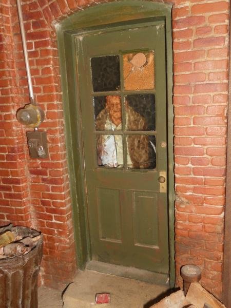 ORIGINAL MICHAEL GARMIN CITY SCAPE SCULPTURE: An elaborate back door scene against a brick wall, the door showing a man peering out, trash and electrical accoutrements all about, lighted, h. 27", w. 15".