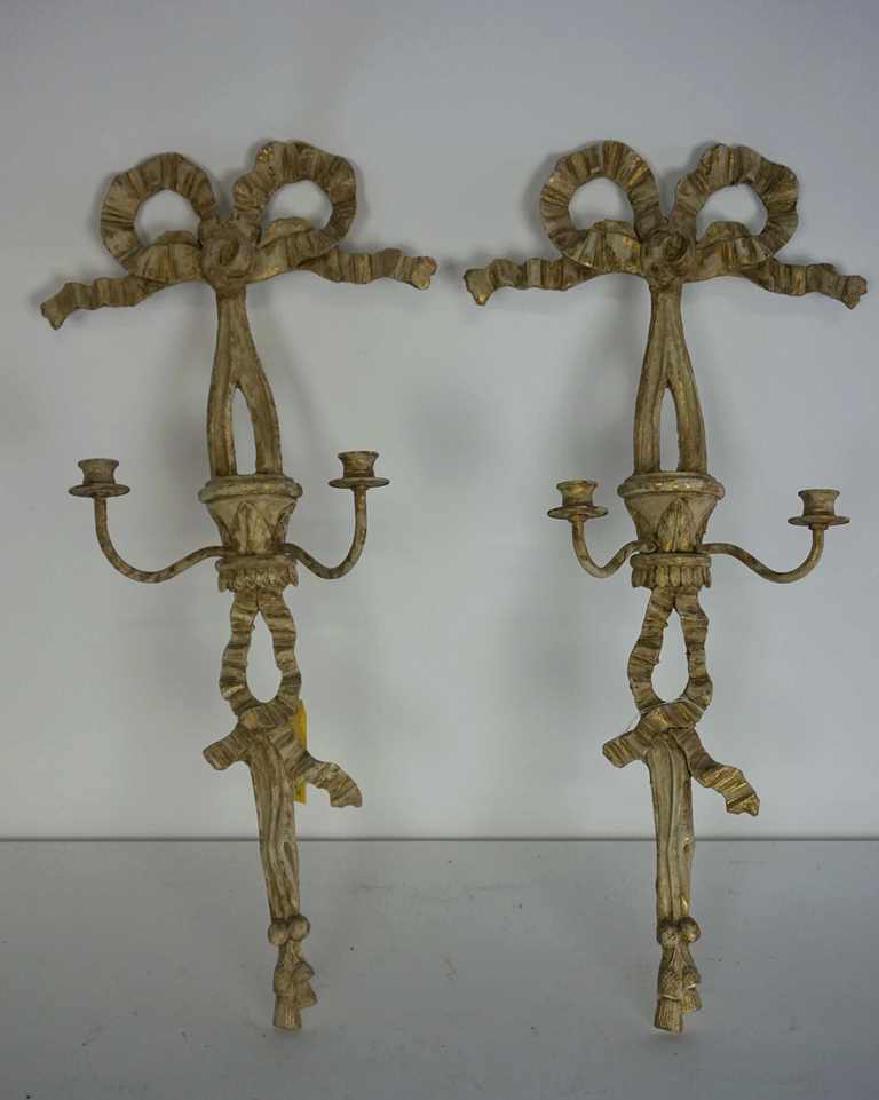 Pair of Carved Candle Sconces: Pair of carved wood candle sconces painted cream and gilt and having a ribbon motif. Two arms to each. 32 inches height, 14.5 inches width, 5.25 inches depth. 32"H x 14.5"W x 5.25"D Measurements: 32"H