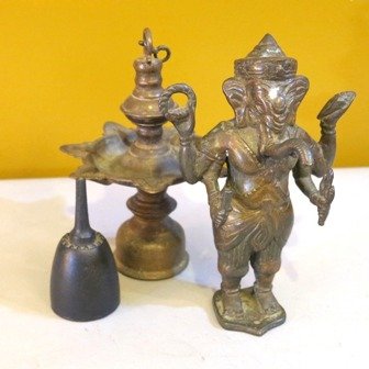 THREE METAL TABLE ARTICLES:: probably Middle Eastern, comprising a bronze standing figure in the image of a god with elephant head, a bell and brass oil lamp. Height of tallest 8 1/2 inches.