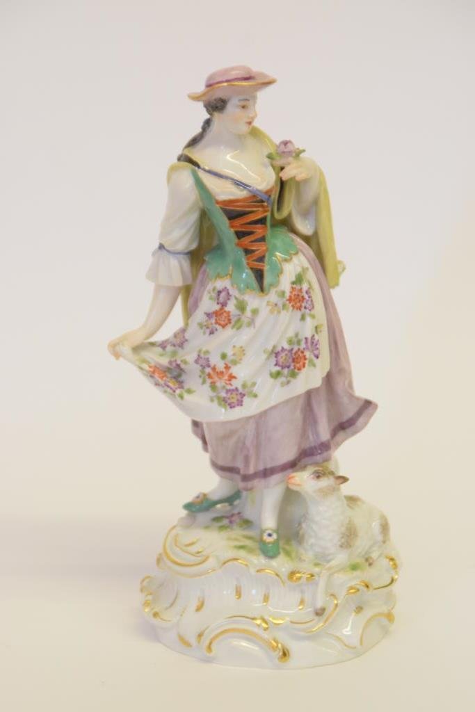 Early 20th Century Meissen Porcelain Figurine: Depicting a woman, a sheep rests at her feet, she wears a floral apron over a purple dress, a cloak accross her shoulders. In her left hand she holds a purple flower. Marked on the back of the figurin