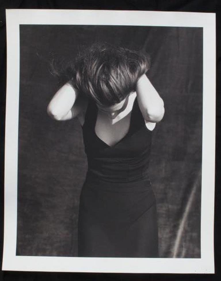 Black and White Print by Michel Comte: Black and white photographic print on baryta paper by Michel Comte. Depicting a female model in a black dress with arms behind her head and her face tilted downward. Artist marks in pencil and stamp t