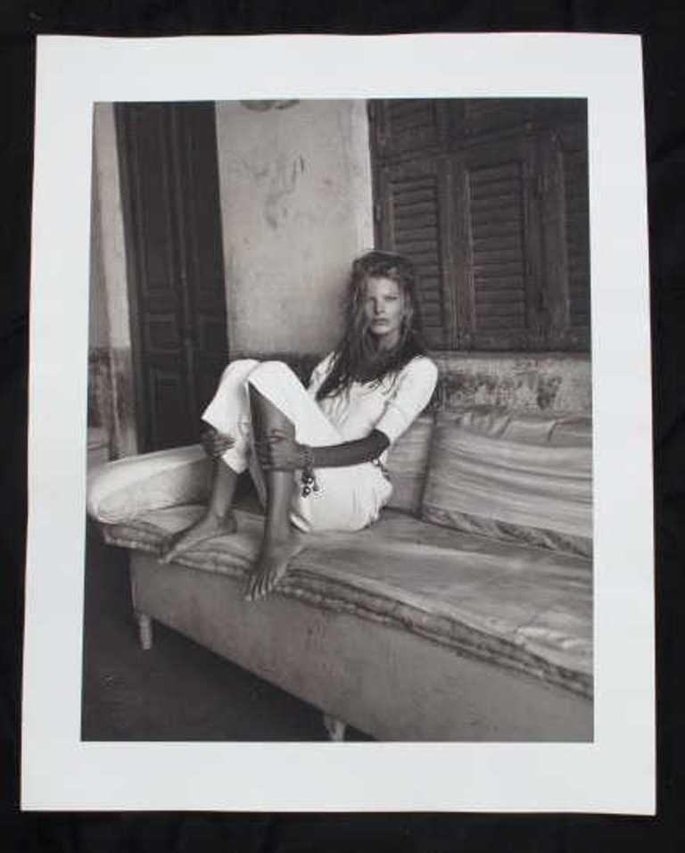 Photographic Print by Michel Comte: Black and white photographic print on baryta paper by Michel Comte. Depicting a female model dressed in white sitting in a rustic interior. Artist's marks in pencil and stamp to verso. 20 inches heigh