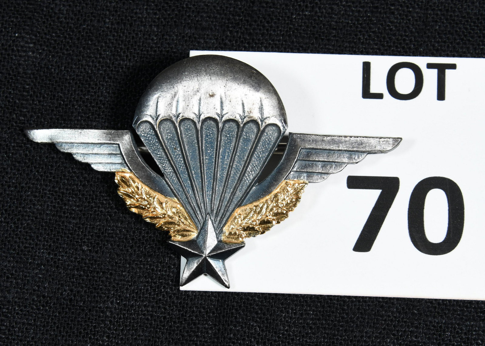 French Airborne Paratrooper Jump Wing Insignia Badge: A French airborne paratrooper jump wing badge. This badge features a parachute with wings above a spray of golden leaves with a star at the center. The reverse side of the pin reads "GS 1185, Drago." 