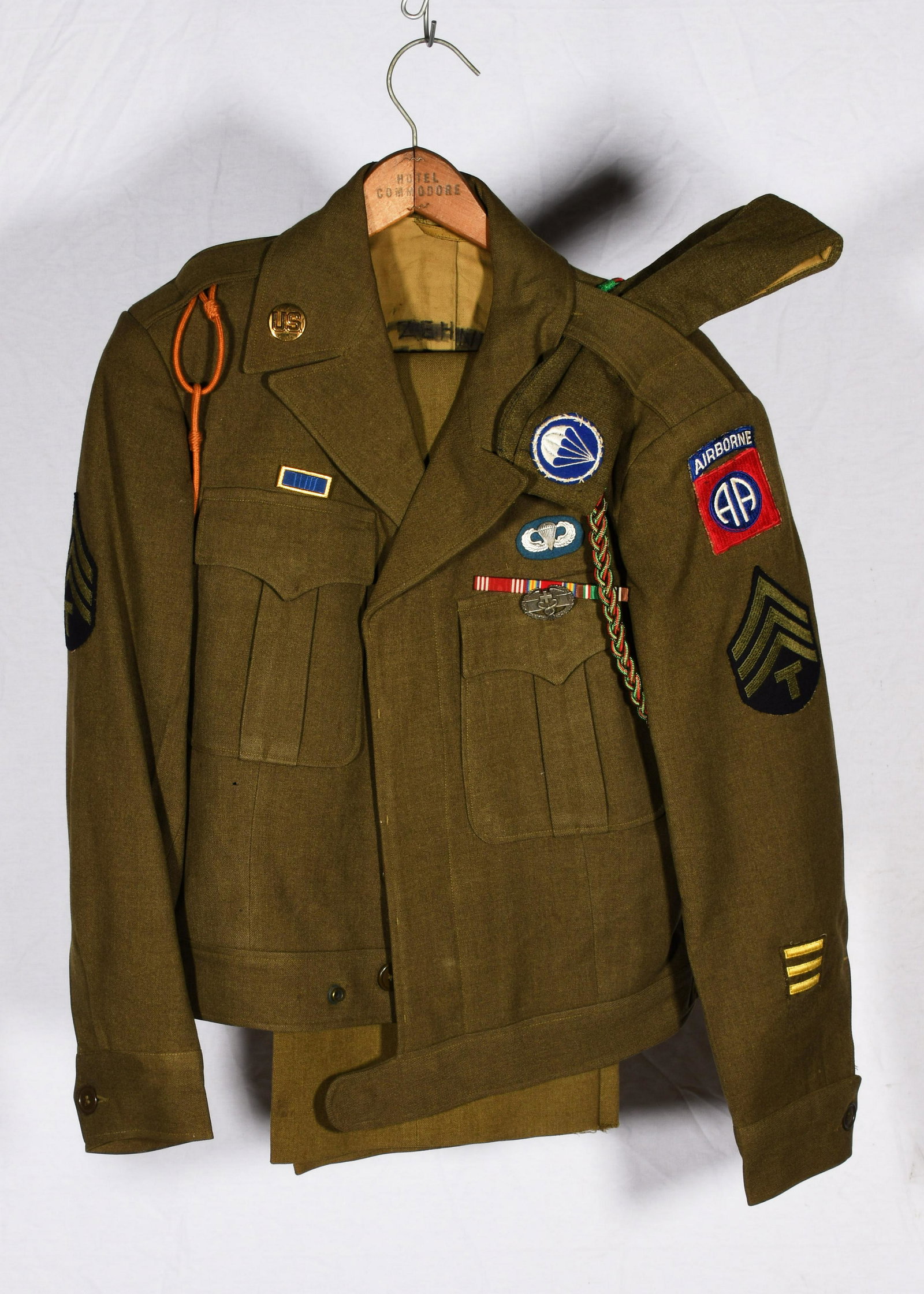 WWII US Airborne Uniform: A US Airborne uniform jacket, pants and hat lot. The jacket has patches for an army technician with 3rd grade stripes. Has a French green and red braided lanyard, an orange lanyard for Netherlands/Hol