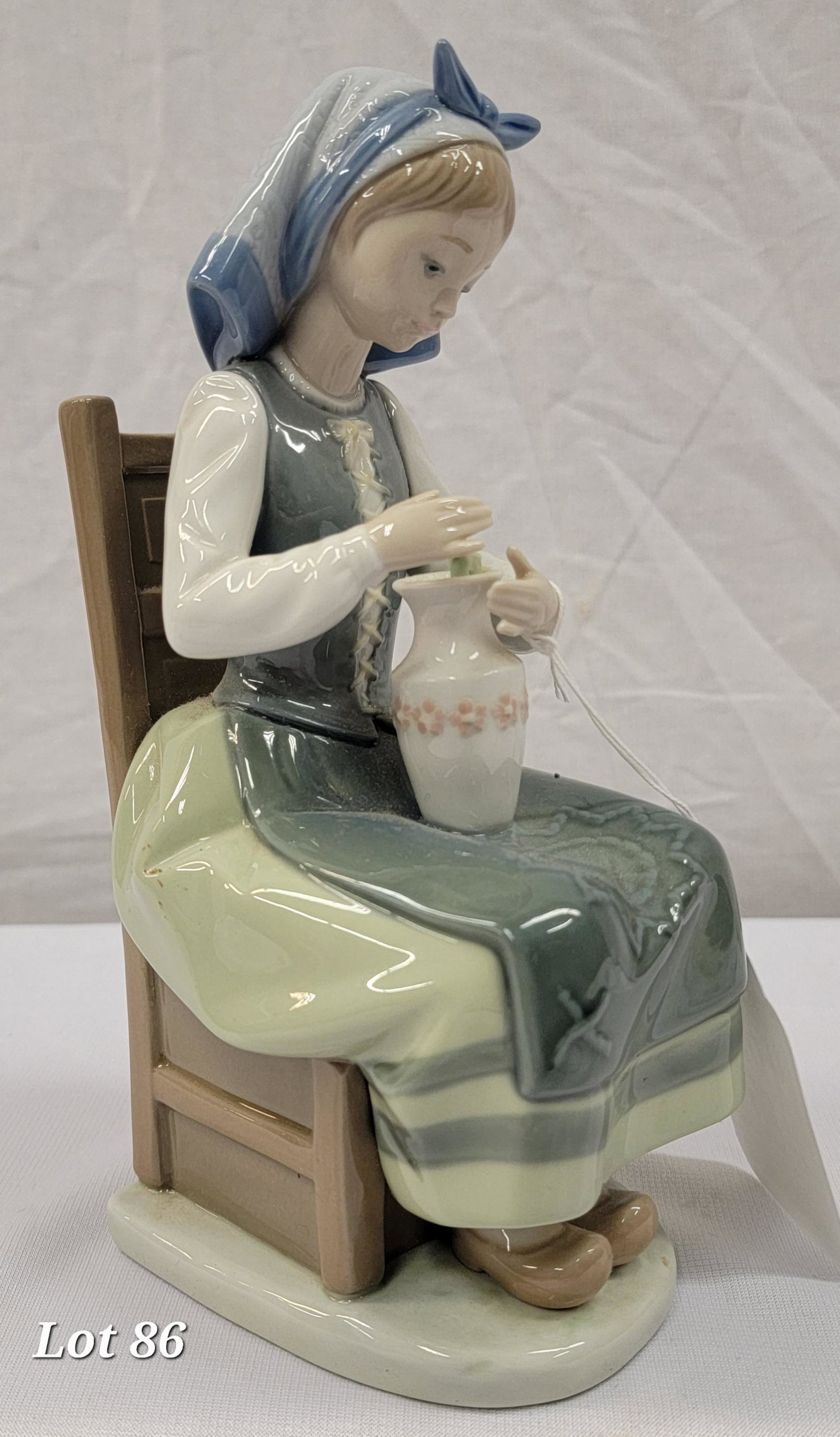 LLadro Porcelain "Flower Harmony" Figurine: A LLadro "Flower Harmony" porcelain figurine. Model no. 1418. This piece features a dutch girl sitting on a chair holding a vase. The flowers have been broken off. The underside of this statue is mark