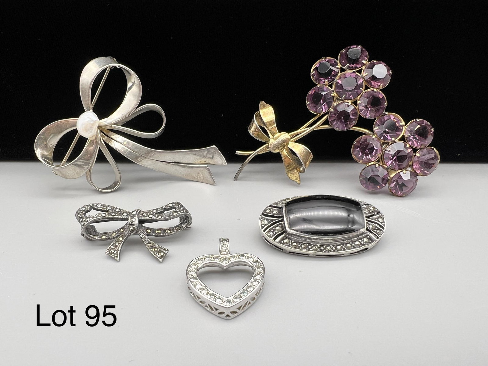 Sterling Lot Including Marquiste: A nice selection of sterling silver jewelry. Includes an amethyst flower brooch, an onyx and marcasite brooch, a heart pendant and two bow brooches.