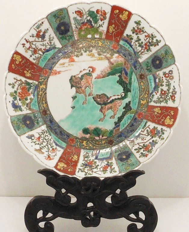 18-19th Century Famille Rose Charger with Stand: China, 2 3/4" tall X 14 1/4" diameter, porcelain charger decorated with bands of floral designs, panels with flowers trees and shrubs, center has two Foo Dogs in a landscape scene, scalloped rim, unma