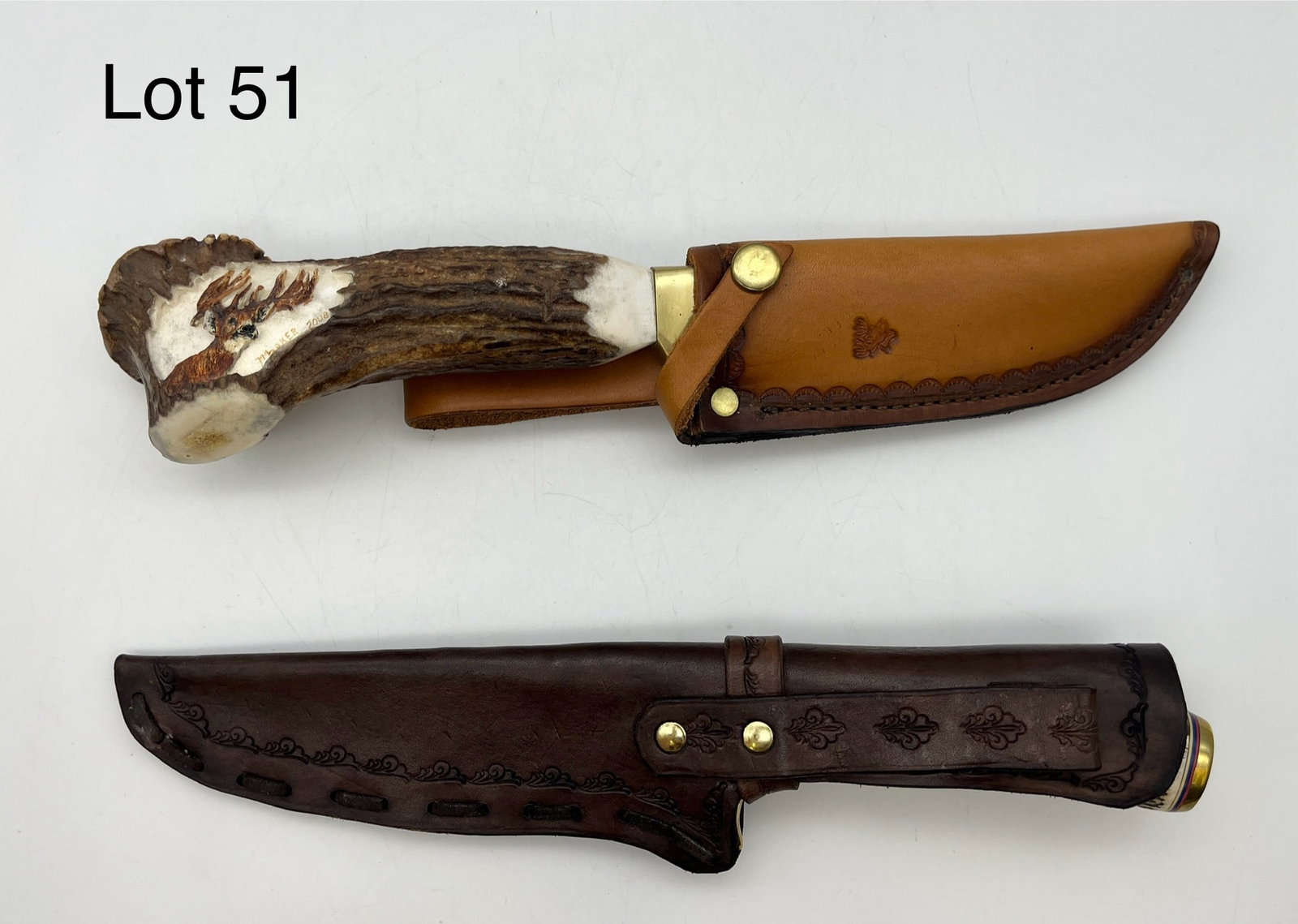Pair of Handmade Antler Knives: A pair of handmade knives with carved antler handles. The first shows a damascus blade in a light brown leather sheath with embossed designs, its handle carved with a portrait of a deer and signed "Mc