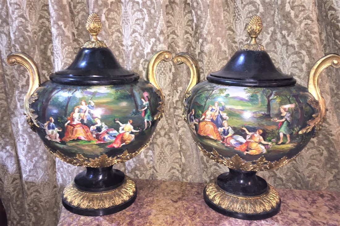 Pair Of Classical Capped Urns.: Highly decorative and painted in the round depicting a lush landscape with gathered figures and musical instruments. The bodies accented with carved gold-gilt foliate overlay. The caps with a berry-cu