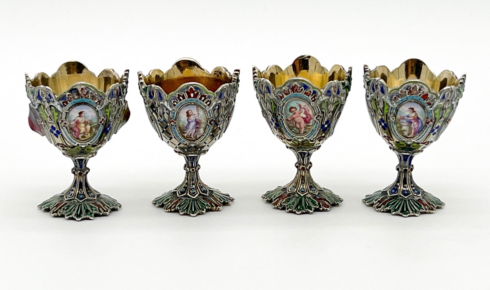 Four Enameled And Silvered Egg Cups: European, 20th Century, Unmarked, Gold Wash Interiors, Reserves Of Trophies And Figures, Height 2.5 Inches, Residue Throughout, Pitting To Interior Of One. See Photos For Additional Condition Informat