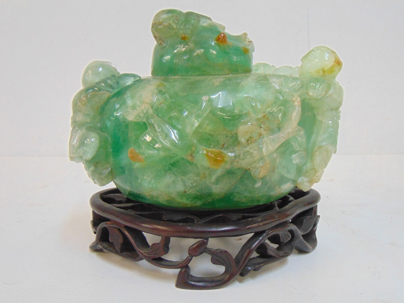 Carved jadeite jar with lid, two figures carved into sides, on wood base, height is7" (as shown): Carved jadeite jar with lid, two figures carved into sides, on wood base, height is7" (as shown)