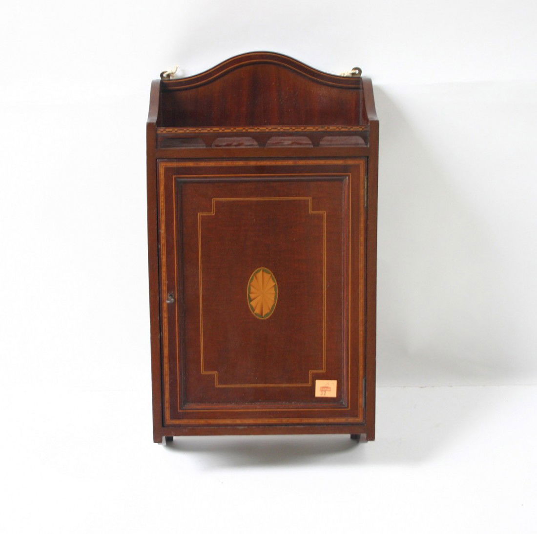 A good quality mahogany Hanging Wall Cupboard, Edwa: A good quality mahogany Hanging Wall Cupboard, Edwardian, with the trade label of John Taylor & Son, Edinburgh, the arched and inlaid back above a pierced gallery and a single panel door enclosing a s