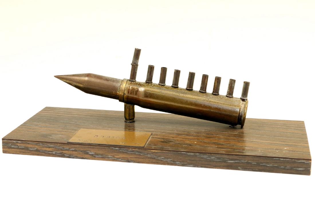 A TRENCH ART MENORAH. Israel, c. 1970. On wood base: A TRENCH ART MENORAH. Israel, c. 1970. On wood base with large caliber main body and 20 caliber oil holders. 7” tall.