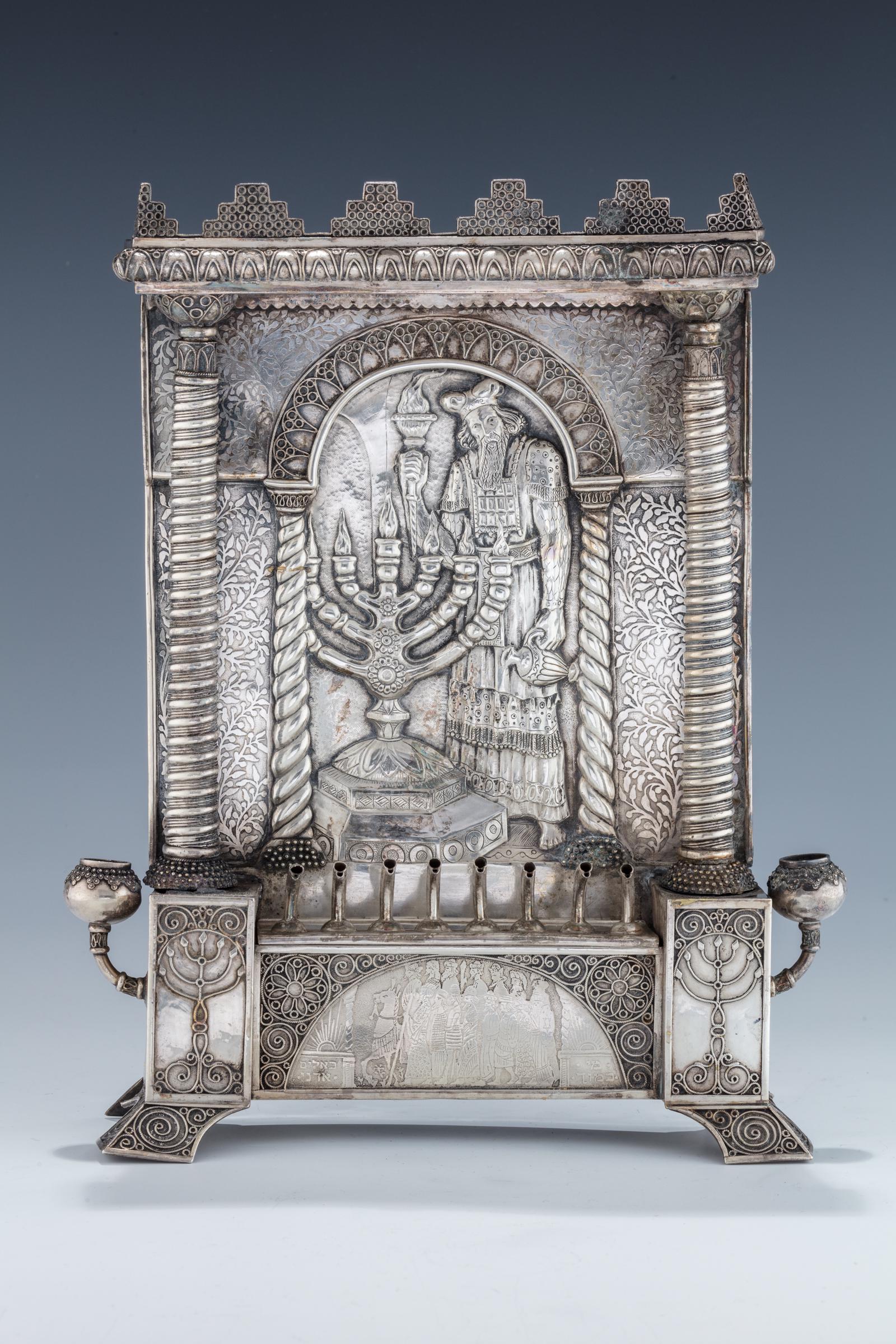 AN EXCEPTIONAL SILVER HANUKKAH LAMP BY BEZALEL.: AN EXCEPTIONAL SILVER HANUKKAH LAMP BY THE BEZALEL SCHOOL. Jerusalem, c. 1920. Portico form. The backplate chased with a towering figure of Aaron the High Priest lighting the menorah in full garb with