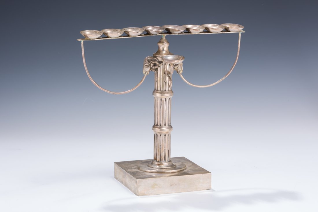 A SILVER MENORAH: A SILVER MENORAH. Germany, c. 1890. On square base. With main Solomonic pillar. Further decorated with two rams’ heads. With original servant lamp. Marked. 9” tall.