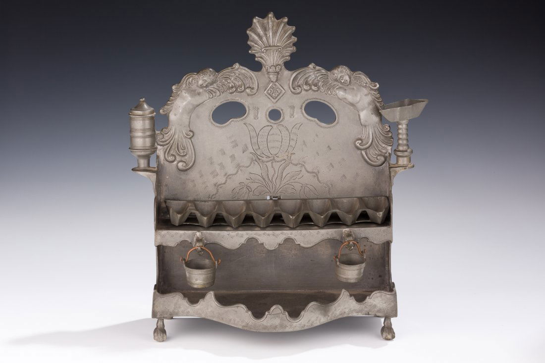 A RARE AND IMPORTANT PEWTER CHANUKAH LAMP. Germany, 18t: A RARE AND IMPORTANT PEWTER CHANUKAH LAMP. Germany, 18th century. On four feet. With two hanging pots to catch dripping oil. Decorated with two cherubs on upper periphery. Marked. 11” tall. Estimate