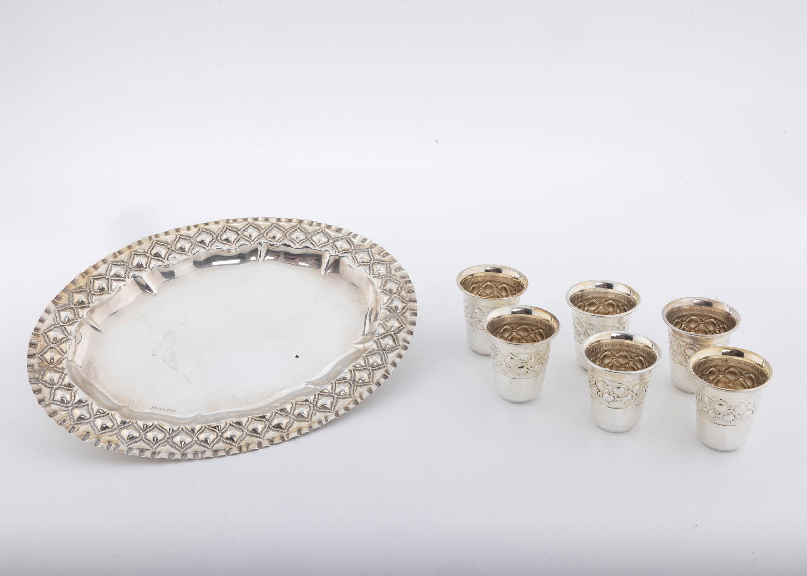 A SET OF SIX STERLING SILVER KIDDUSH BEAKERS ON A TRAY BY HADAD BROTHERS.: Israel, modern. Oval form with embossed border. The tray is 8.9" x 6.1" and the cups are 1.7" tall each. 203.5 grams total weight.