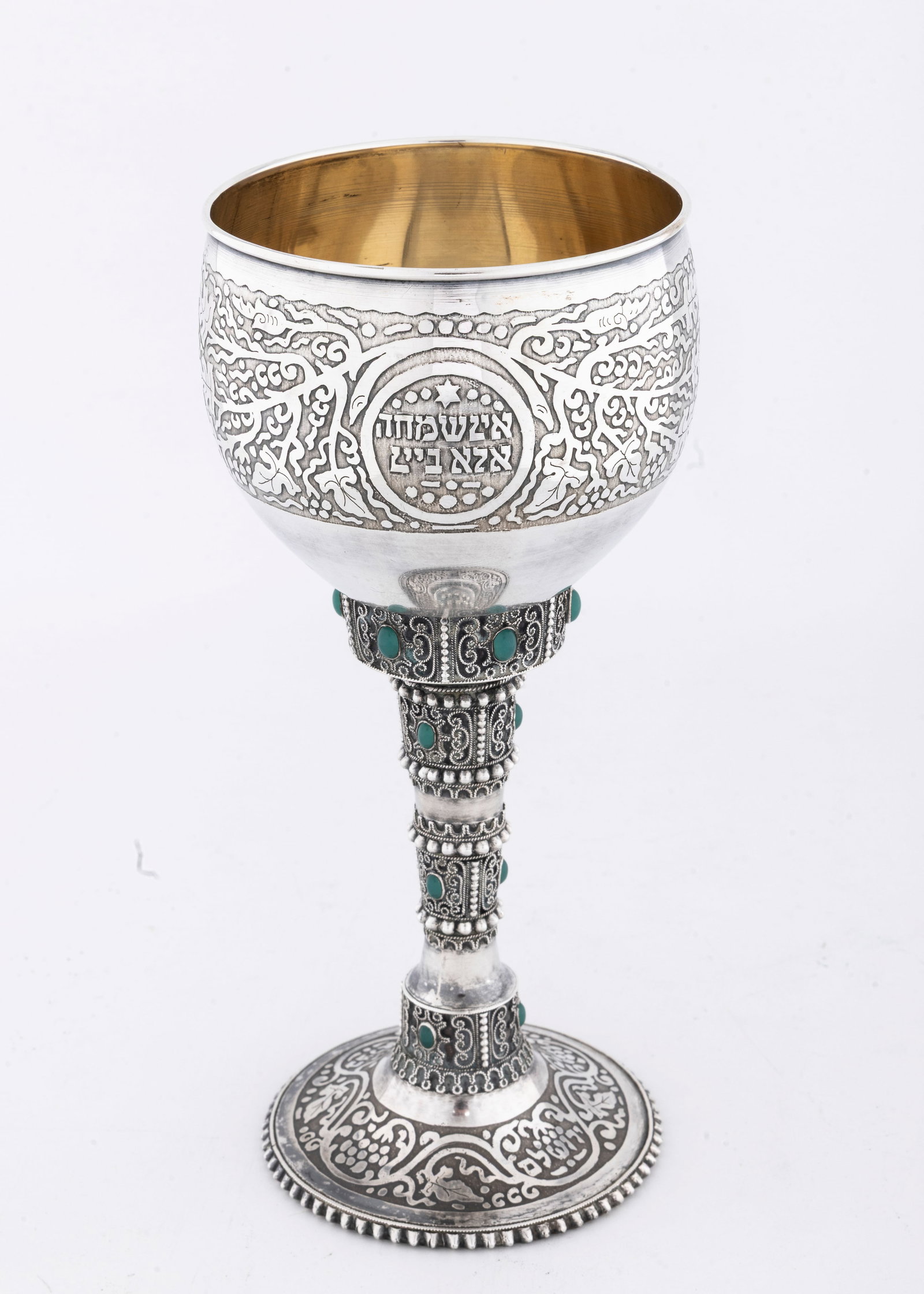 A MONUMENTAL AND EARLY SILVER KIDDUSH GOBLET BY THE BEZALEL SCHOOL. Jerusalem, c. 1920.: On a round beaded basew that is acid etched design with five layer stem decorated with applied wirework and fitted with green stones. The upper portion highly decorated in acid etched design spelling 