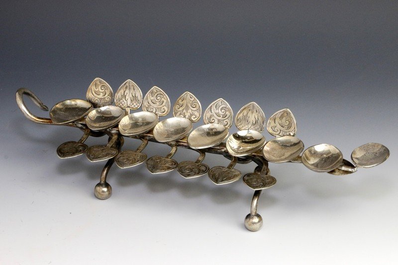 A FOLK ART SILVER MENORAH. Continental, Early 20th: A FOLK ART SILVER MENORAH. Continental, Early 20th century. On four balled feet. With eight spoon shaped oil holders and leaf decoration. Hand wrought. 9” long. Estimate $1,200 – 1,500