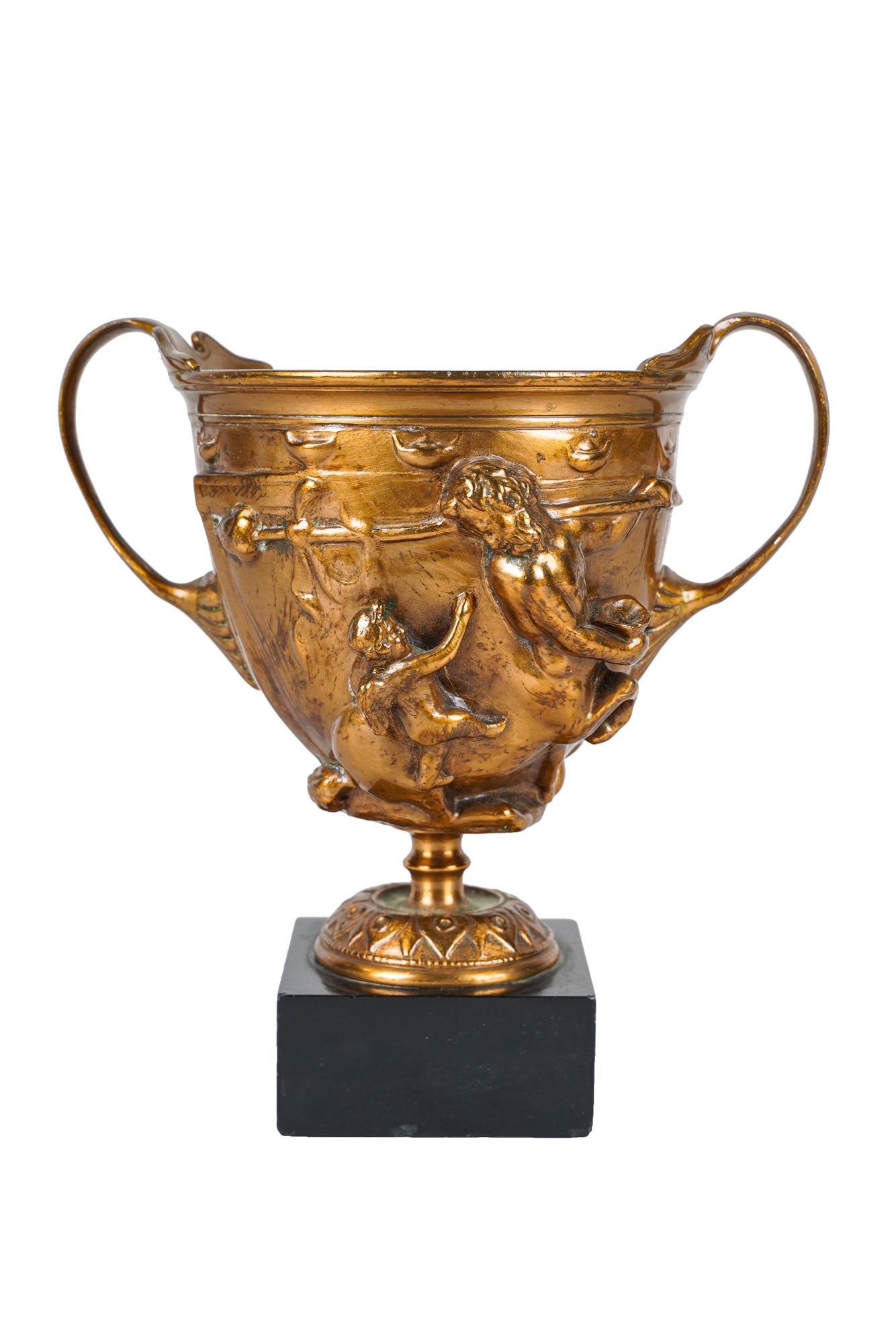 GILT-BRONZE CHALICE: decorated with centaurs and putti, mounted to a black stone base, with liner Condition: with some rubbed wear to gilding. flea bite chips to stone. chalice measuring: 5 1/2 inches: total height: 6 1/2