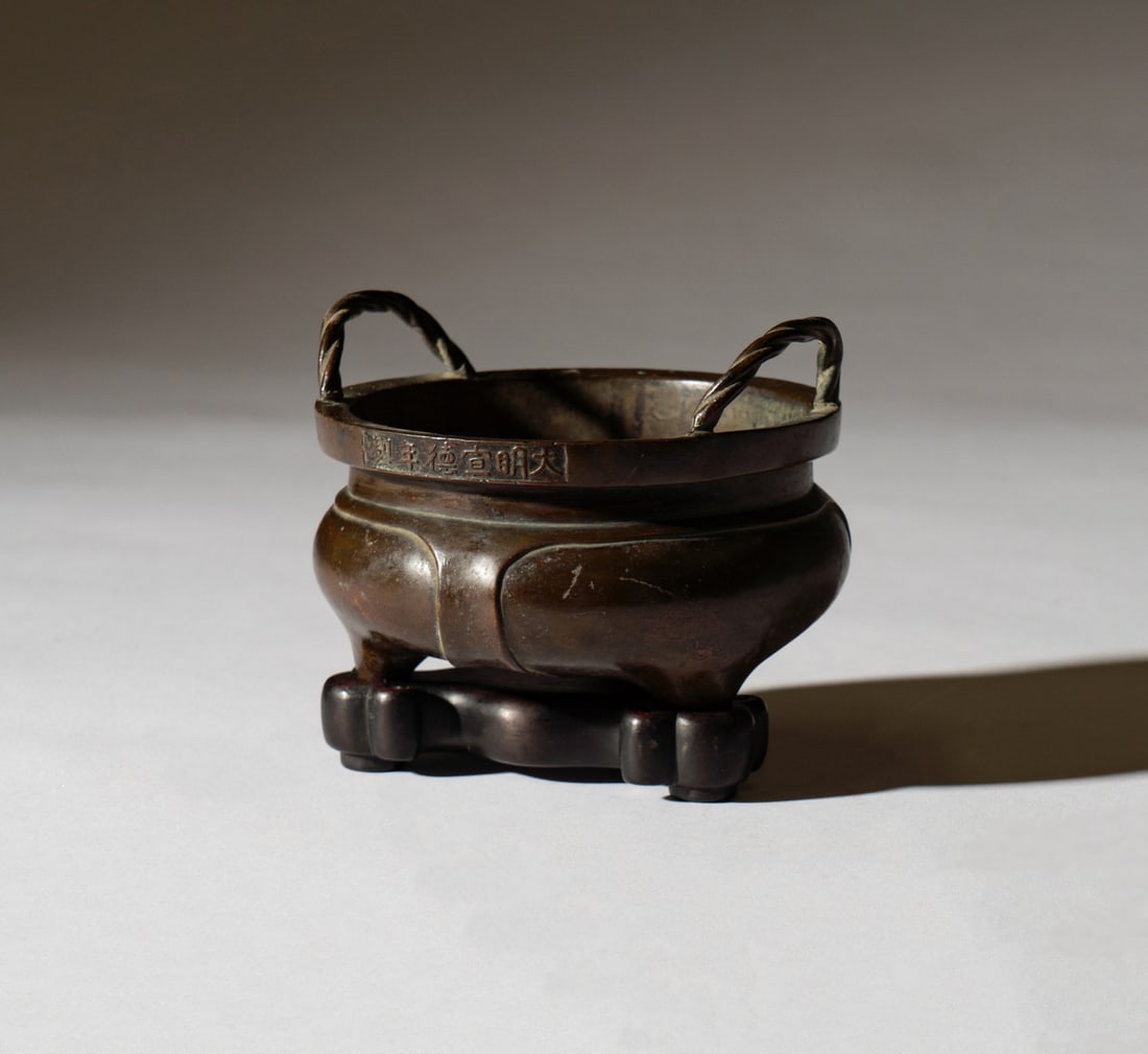 A Chinese Bronze Censer: Ming Dynasty; round with two upright twisted loop handles, raised on three feet, with a wooden stand; inscribed to mouth rim on each side Excluding stand: 3 1/4 x 4 1/2 in. (8.3 x 11.4 cm.)