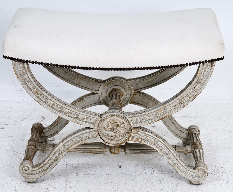 CARVED & PAINTED CURULE-FORM BENCH: early 20th century, in the Louis XVI taste the white upholstered rectangular seat on foliate carved and white painted curule supports centering on a round floral carved medallion and having scrolling