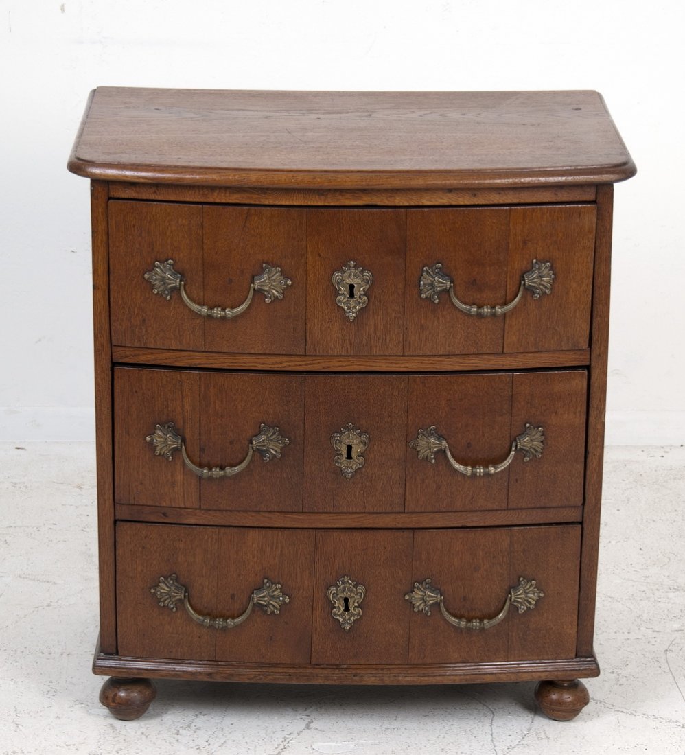 FRENCH PROVINCIAL STYLE OAK COMMODE: the top having a gently bowed front edge above a case of three drawers, each with two bronze loop pulls flanking a cartouche escutcheon, on bun feet 29 inches long; 20 inches deep; 31 1/2 inches high
