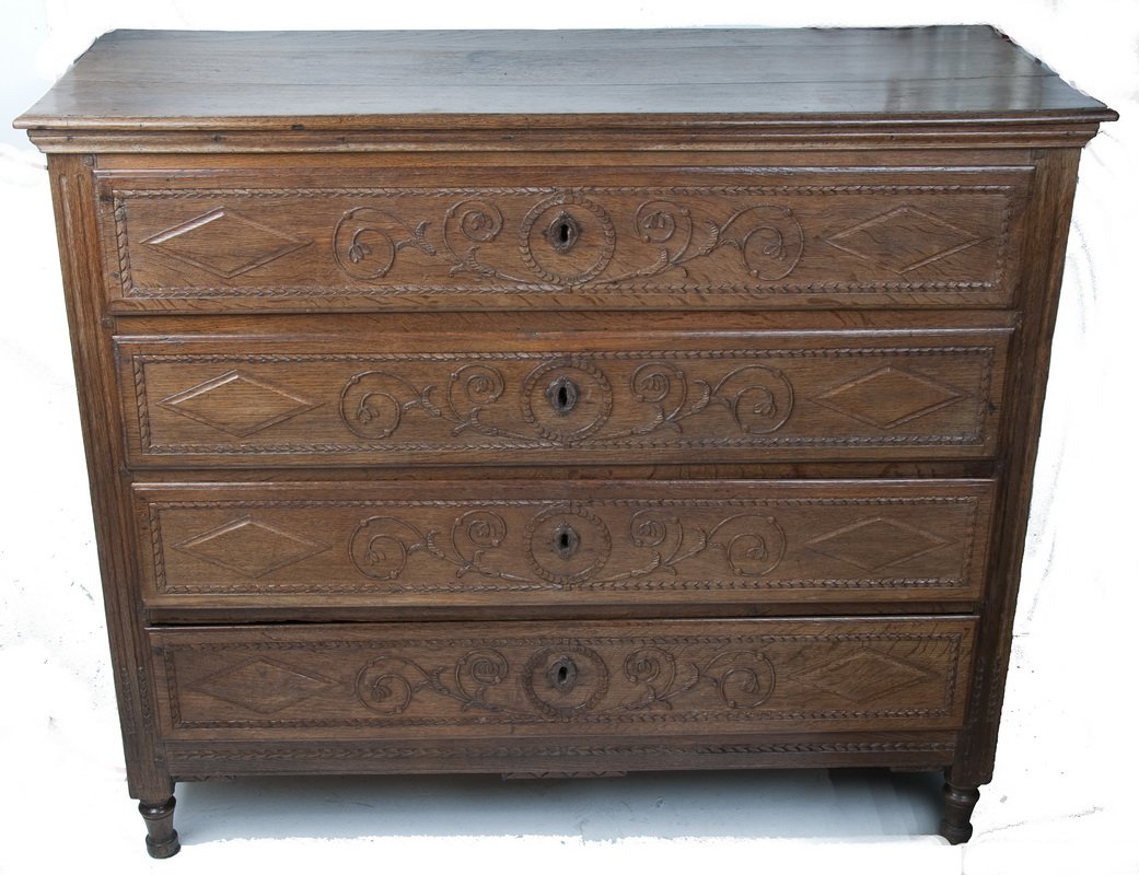 LOUIS XVI STYLE PROVINCIAL OAK CHEST OF DRAWERS: having a rectangular top and a case of four long drawers, each carved with a laurel border around two rhombuses flanking a floral design and wood escutcheon; on bun feet Provenance: The Estate of Davi