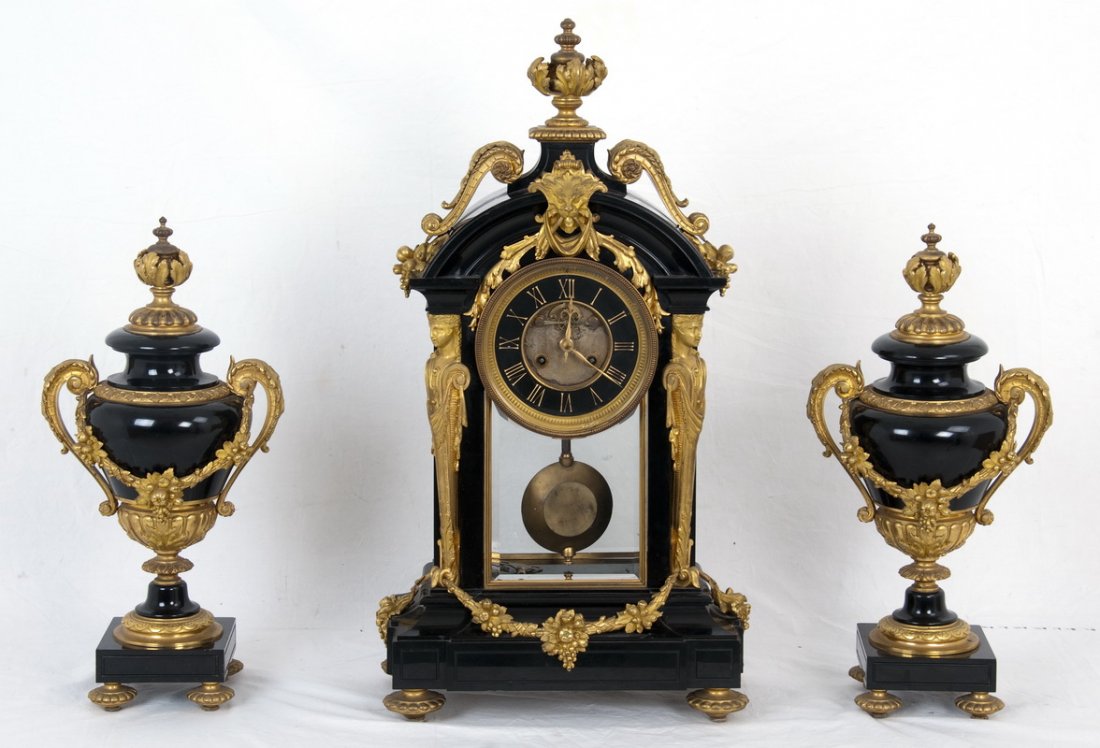 THREE-PIECE FRENCH ORMOLU-MOUNTED MARBLE CLOCK SET: the black marble clock having a curved pediment with a gilt bronze urn finial, scroll, masque, and swag mounts, and a pair of caryatids flanking the round dial and glass door; each urn of a similar fa