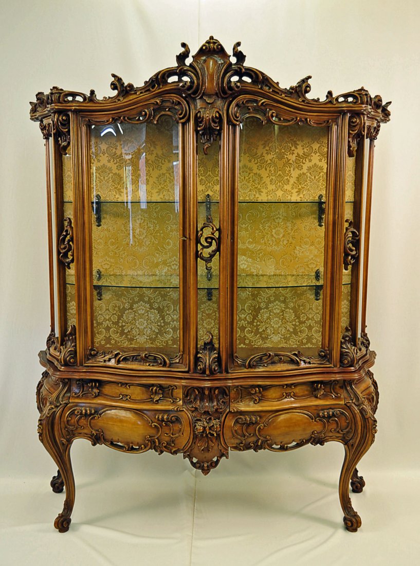 French Rococo Style Vitrine with Curved Glass: French Rococo style hand carved two part vitrine, last half 20th Century with curved glass and glass shelves. Four drawers below two curved glass doors. Back of interior is covered with gold floral ma
