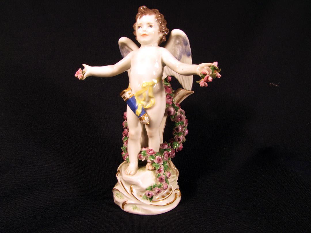 Joyful Meissen Cherub Figurine: 7 1/4" standing cherub with his quiver tethered at his waist. A floral garland laces the tree trunk and the garland that he should be holding is missing, instead he is holding only the ends in his han
