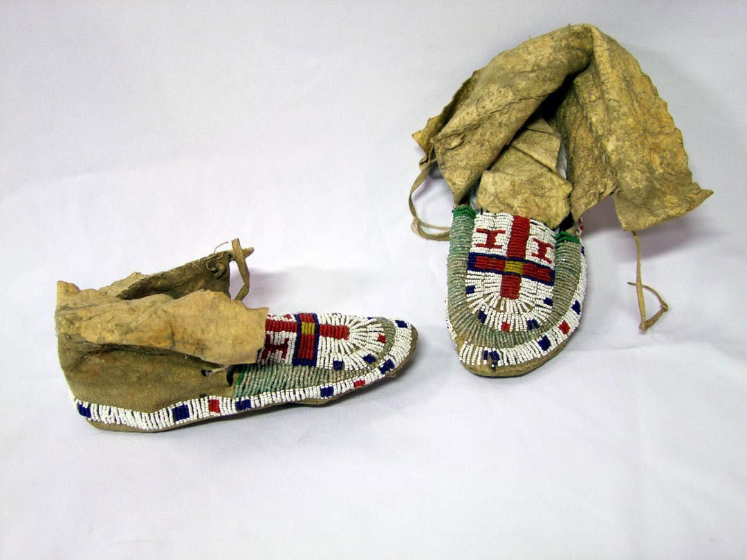 Beaded Indian Moccasins: Sinew stitched moccasins with hard leather soles, beaded with red, white, blue and two shades of pale green glass beads. Greasy yellow beads in the center of the cross. 8 1/2" long. Three rows of whit