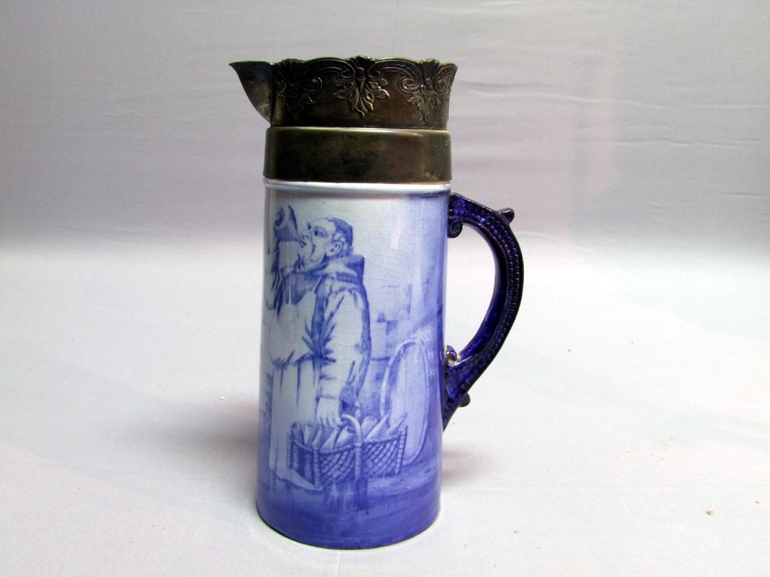 Flo Blue Tankard: Decorated tankard showing a jolly Monk sampling his wine. The silver plate top is worn. Shows crazing. 11 1/8" high. No chips, cracks nor repairs