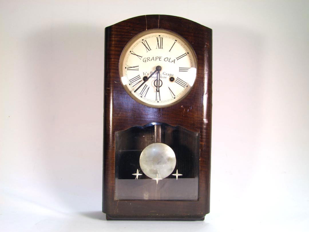 GRAPE OLA Advertising Clock: Wall clock with advertising on the face, "Grape Ola, It's Real Grape, Sold Here". 9 1/2" x 4 1/2" x 18 1/2" high. Veneer patch on upper front.