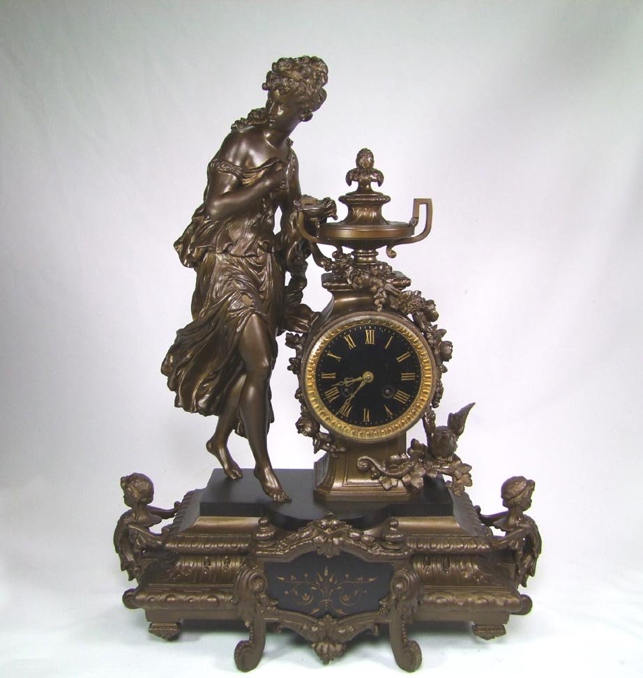 1880s Figural Clock: Spelter and slate clock with Grecian woman holding a betty lamp and standing on black slate. Floral swags drape around the clock with a bird perched on one side. Winged children form the side feet. De