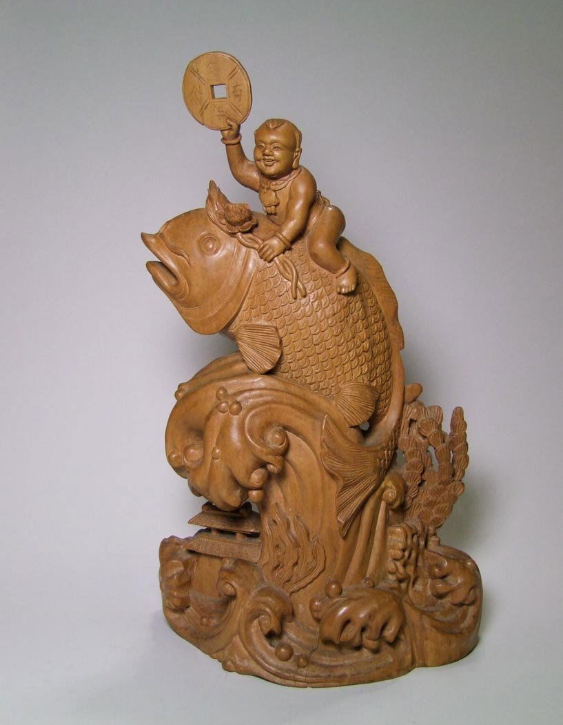 Chinese Wood Carving: 15" wood carving of a happy boy riding the back of a fish as it is carried out of the water upon a large wave. The happy boy holds a coin in one hand and two stems of a lotus in the other. A small pag