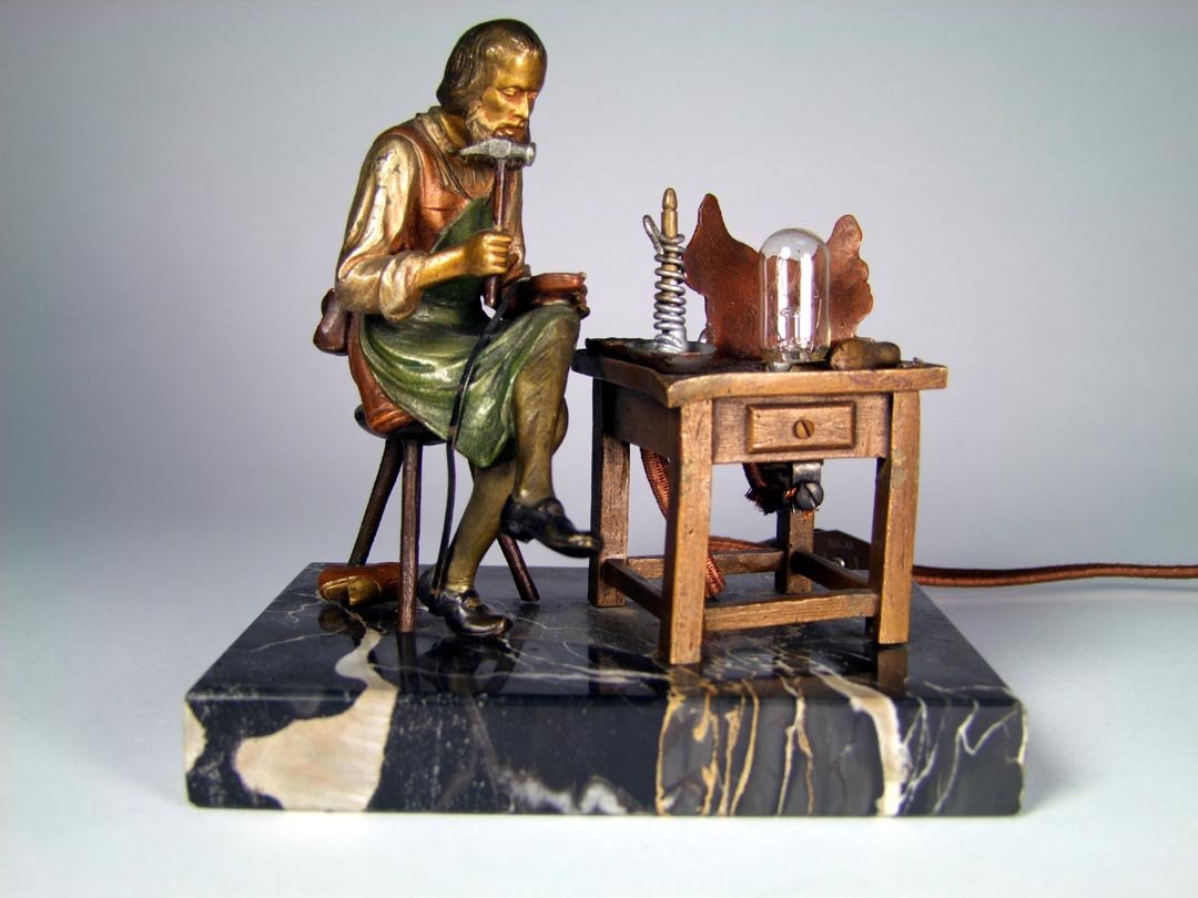 Austrian Bronze Lamp: Cold painted bronze of a cobbler sitting at his work bench working on a shoe. On the top of his work table is a candle, a bowl, shoe form, cluster of tacks and a piece of leather that is draws up to r