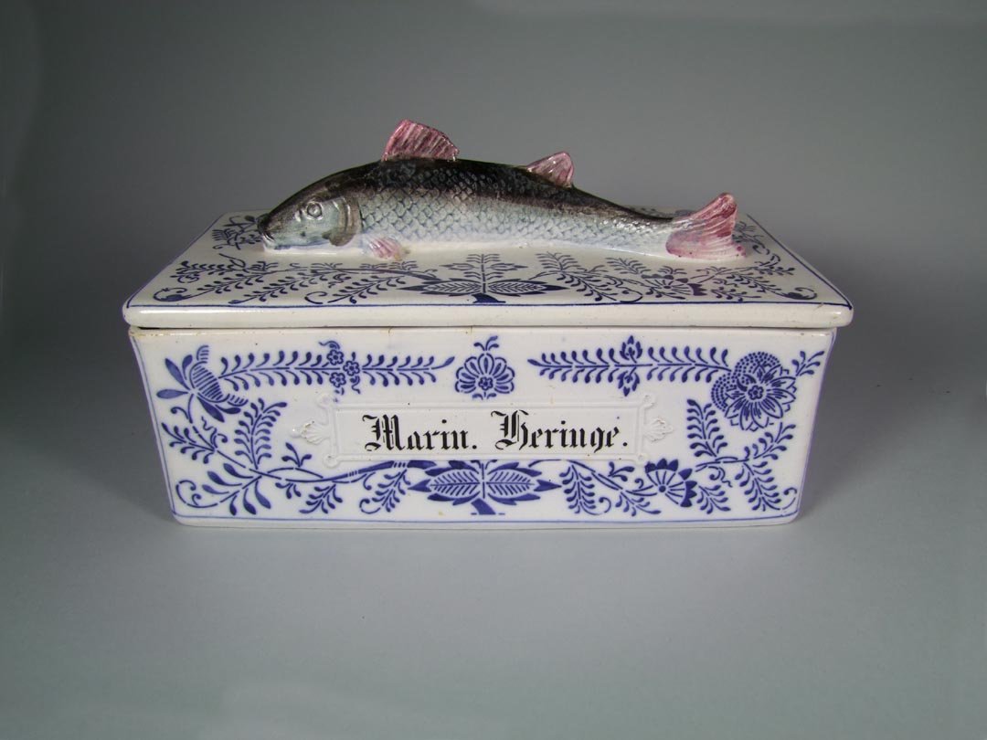 Sardine Box: Blue and white porcelain box with a fish forming the handle on the lid. "Marin Heringe" is written on the front of the box. Decorated in the blue onion style.13 1/4" x 6" x 6 3/4" high. A few small gl