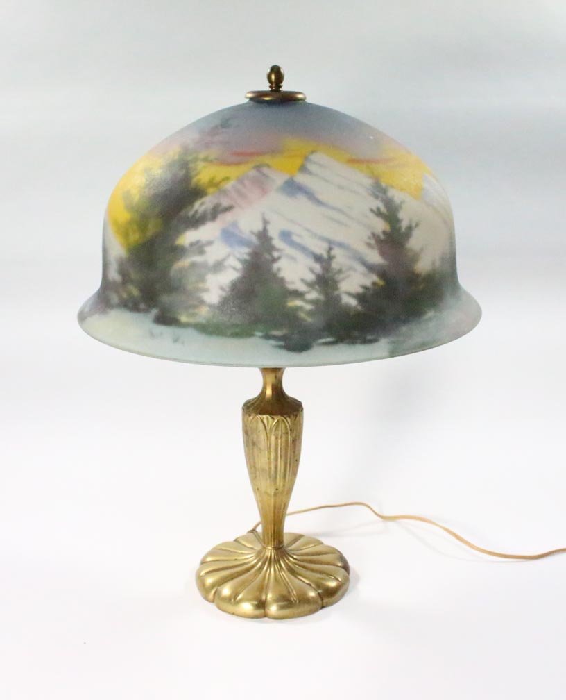 Reverse Painted Pairpoint Lamp: Signed bell-shape reverse painted shade with pine trees against a mountainous background. 13 3/4" diameter x 8" high. Signed Pairpoint gold tone base with two sockets 19" high. One small chip on the s