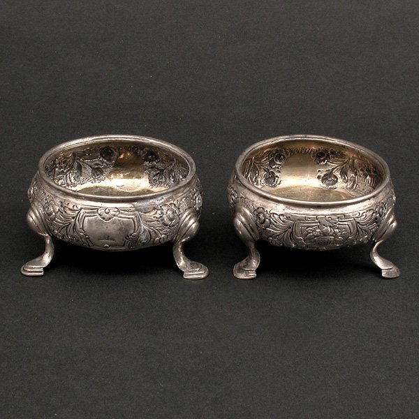 British Sterling Salt Cellars, 1737 & 1748: CELLAR. Two British sterling silver salt cellars, one hallmarked 1737 the other 1748, each with a with a bowl decorated in repousse floral baskets and scrolling flowers, set on three curved legs on pa