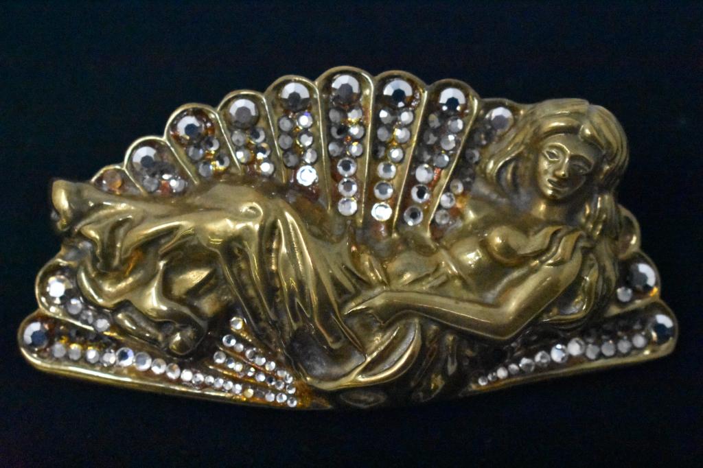 Ceasar Ugarte Nude Art Production Belt Buckle: Topless lady goddess in the Art Nouveau style, is reclining surrounded by rhinestones inlaid in a fan. Belt buckle appears to be brass and all rhinestones are present. 5” long.