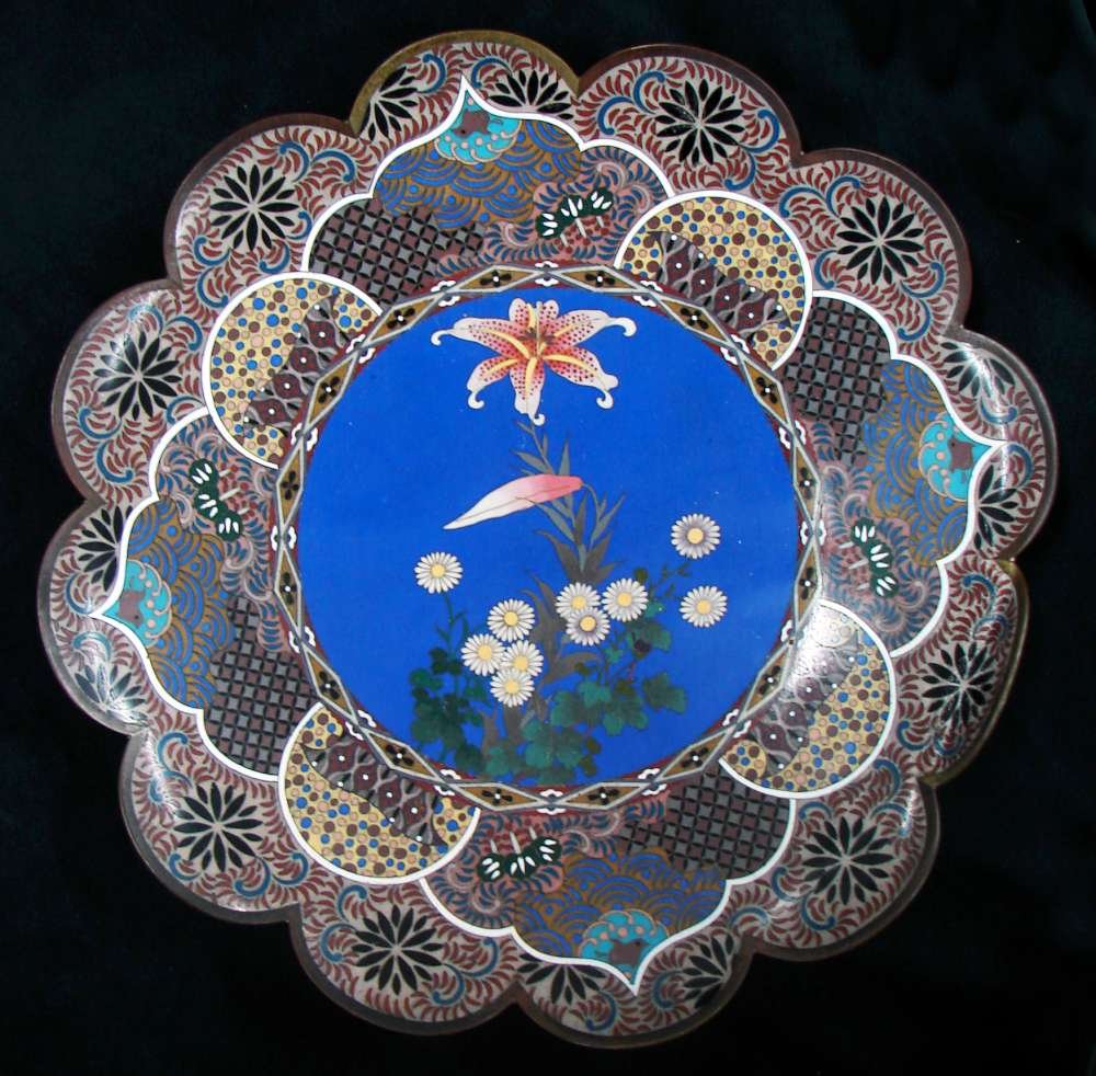 Complex Meiji Period Circa 1890 Japanese Lily Char: Lily Charger with Japanese Cloisonne enamel. Daisy flowers and elaborately patterned edges. One edge flap is bent and the enamel on both sides is damaged. 2" height by 14 1/2" diameter.