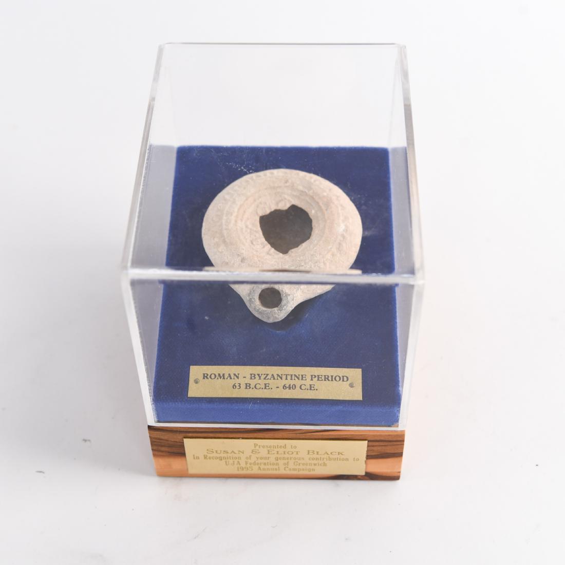 ANCIENT ROMAN OIL LAMP: Dedicated recognition plaque on display case. Marked as "Roman - Byzantine Period 63 BCE-640 CE." Dimensions: (Case) H 5.25" x W 4.25" x D 5.25", (Vessel) H 1" x Dia. 3.25" Condition: Wear consistent 