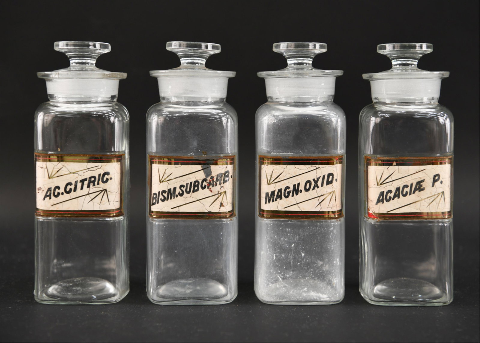 ANTIQUE LABEL UNDER GLASS APOTHECARY BOTTLES: 4 glass bottles. Dimensions: H 8" x W 3" Condition: Losses and cracks to glass label covering. Small chips to tops.