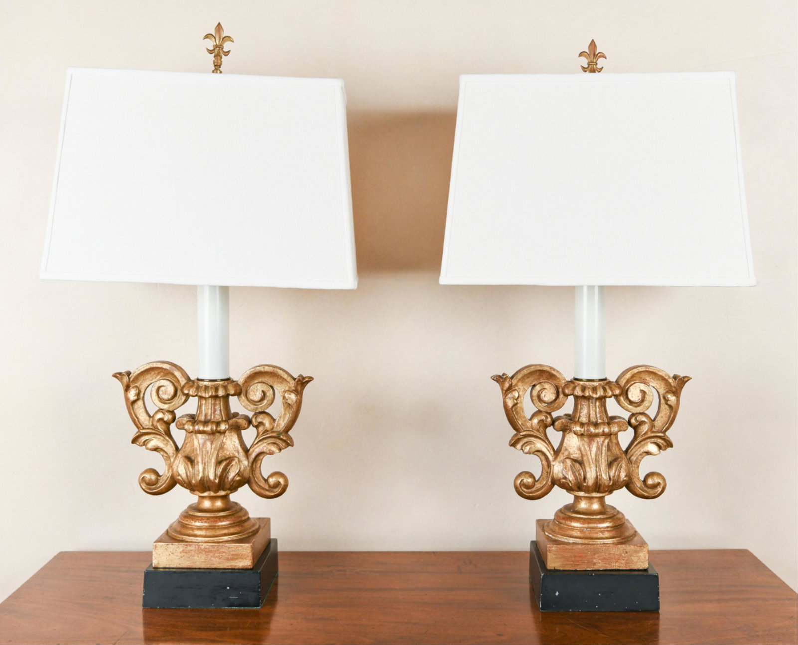 PAIR OF VINTAGE CARVED & FAUX GILT LAMPS: Fleur de lis finials. Carved wood with faux antiqued gilding. Dimensions: (Incl. shade) H 31.25" x W 16.25" x D 10" Condition: One crooked on base. Some wear to gilding. One with break repair to one t
