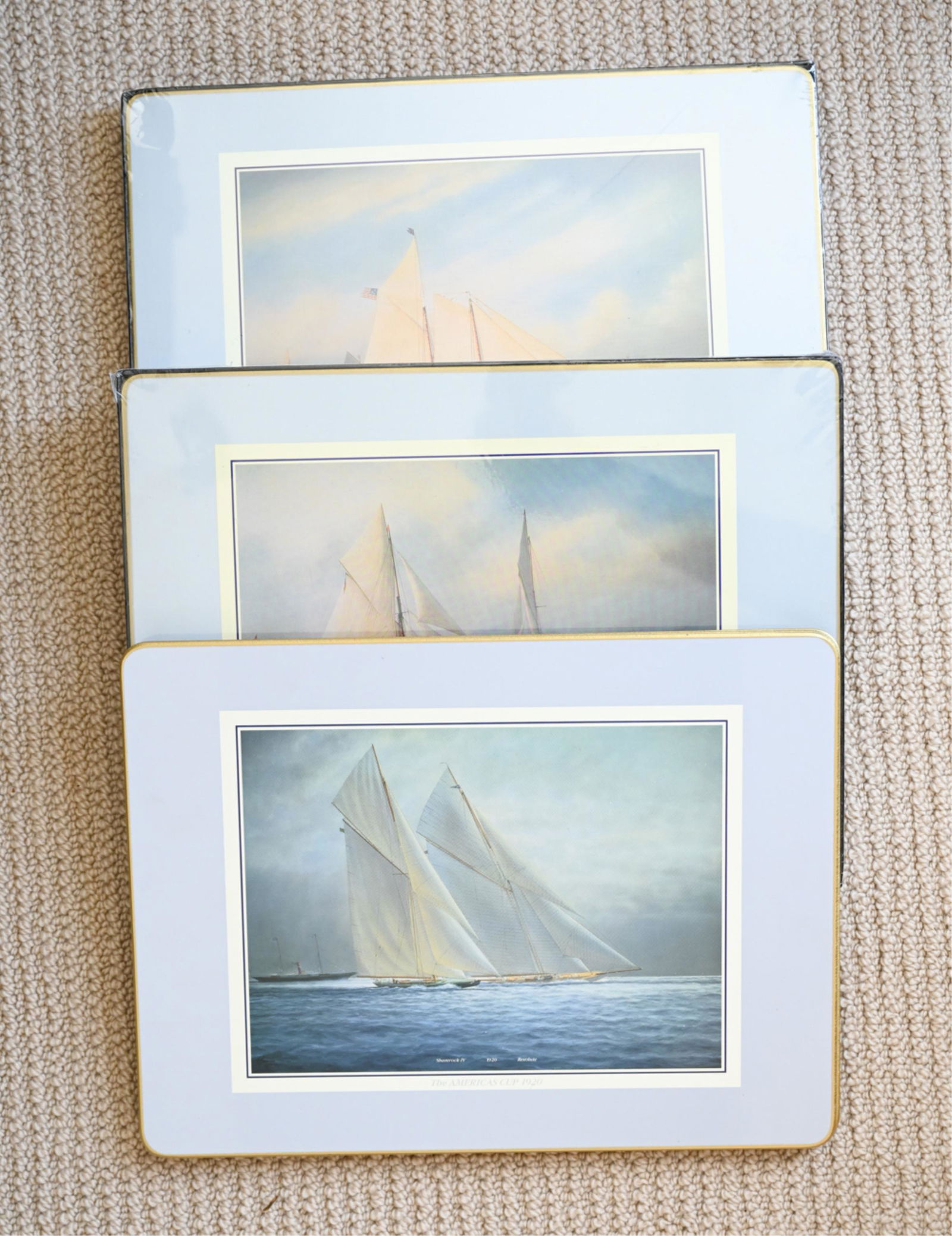 SET OF (12) CLOVERLEAF AMERICA'S CUP PLACEMATS: Laminate with cork backs and gold-painted beveled edges, decorated with various images of yachts. Dimensions: L 11.5" x W 15.5" Condition: Two sets are new in box; the remaining (4) placemats show lig