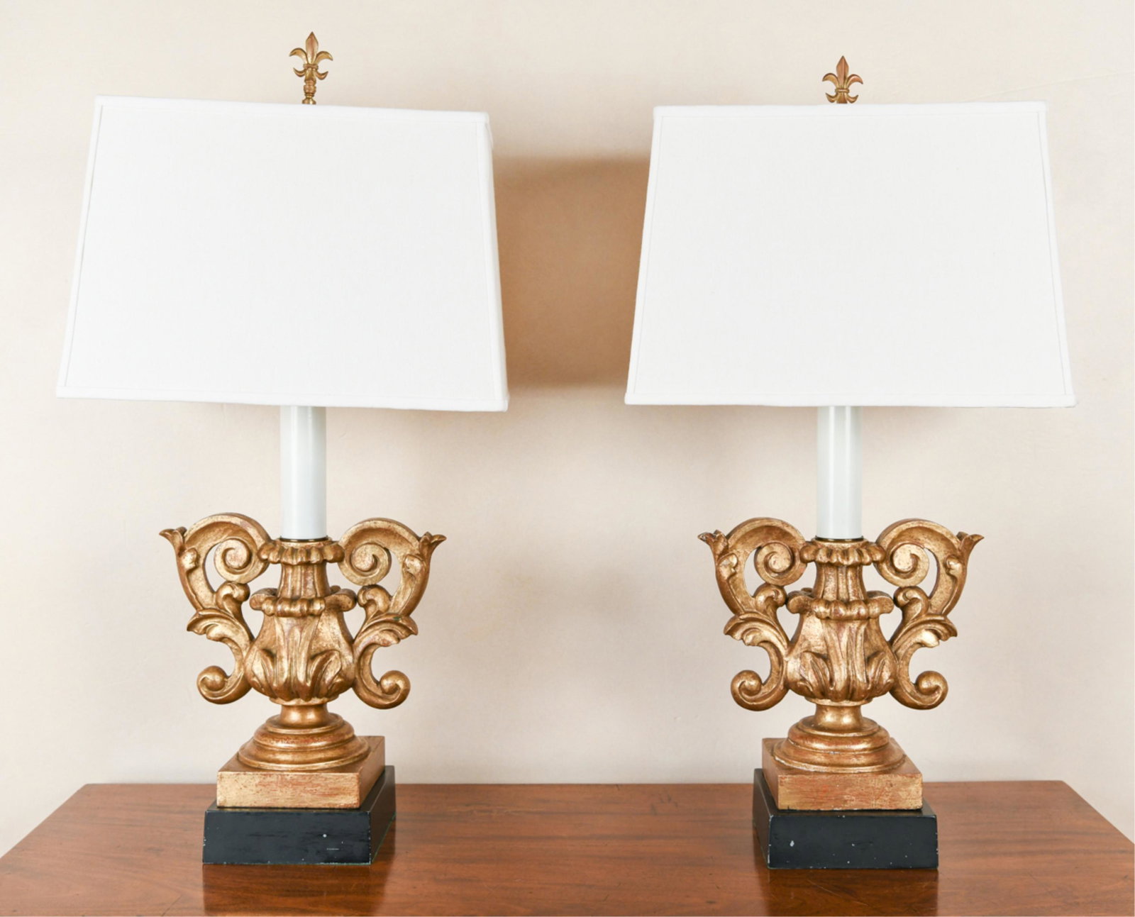 PAIR OF VINTAGE CARVED & FAUX GILT LAMPS: Fleur de lis finials. Carved wood with faux antiqued gilding. Dimensions: (Incl. shade) H 31.25" x W 16.25" x D 10" Condition: One crooked on base. Some wear to gilding. One with break repair to one t