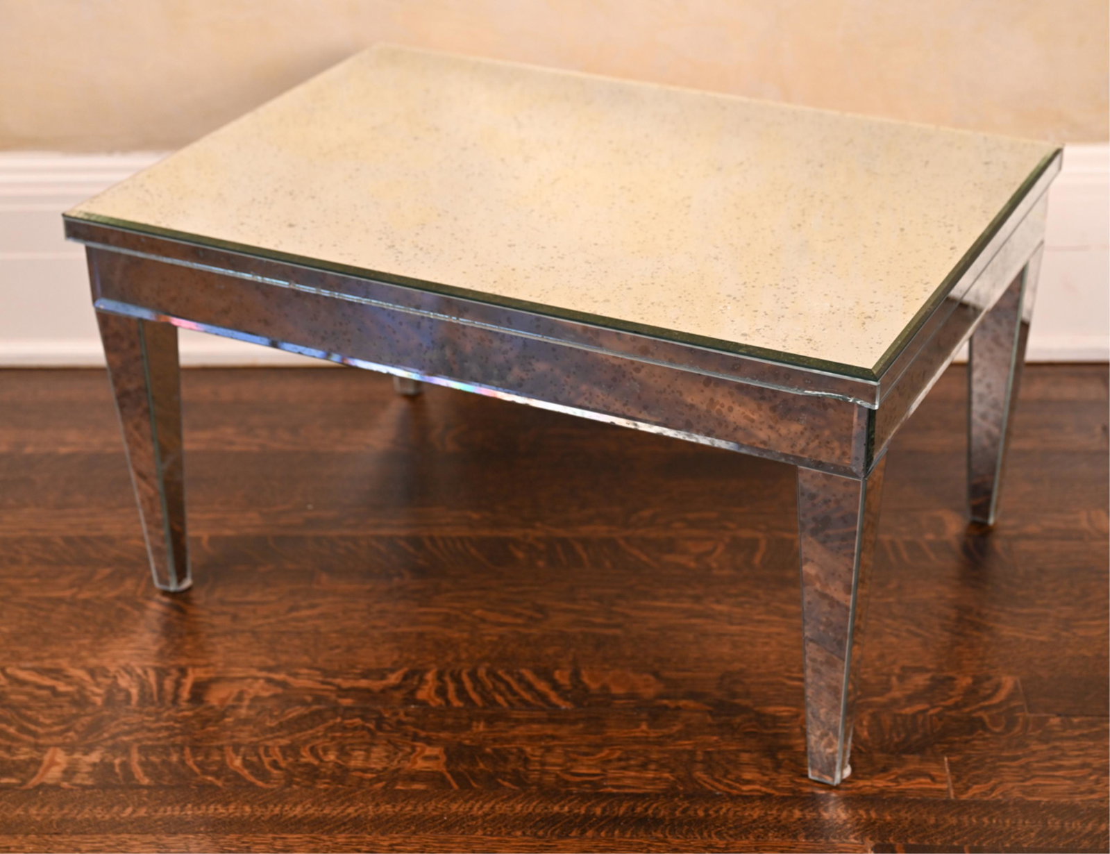 MIRRORED COFFEE TABLE: With beveled edge. Intentionally foxed mirror. Dimensions: H 15.75" x W 29" x D 20.75" Condition: Mirror cracked at one foot. Few small chips to edges. Light wear consistent with use.