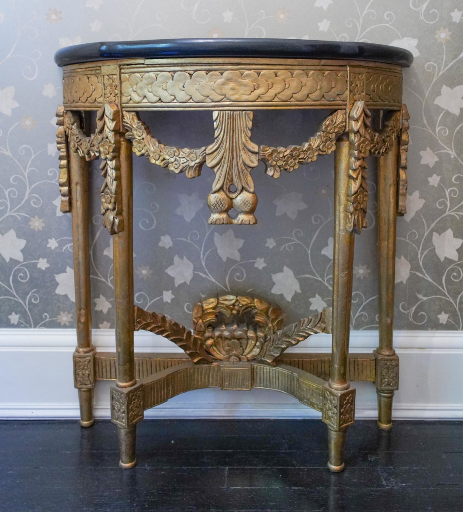 LOUIS XVI-STYLE GILTWOOD & GRANITE DEMILUNE TABLE: Carved and applied giltwood table with elaborate floral garland motifs and black granite top. Dimensions: H 34.5" x W 32" x D 12.5" Condition: Showing light wear consistent with age and use including