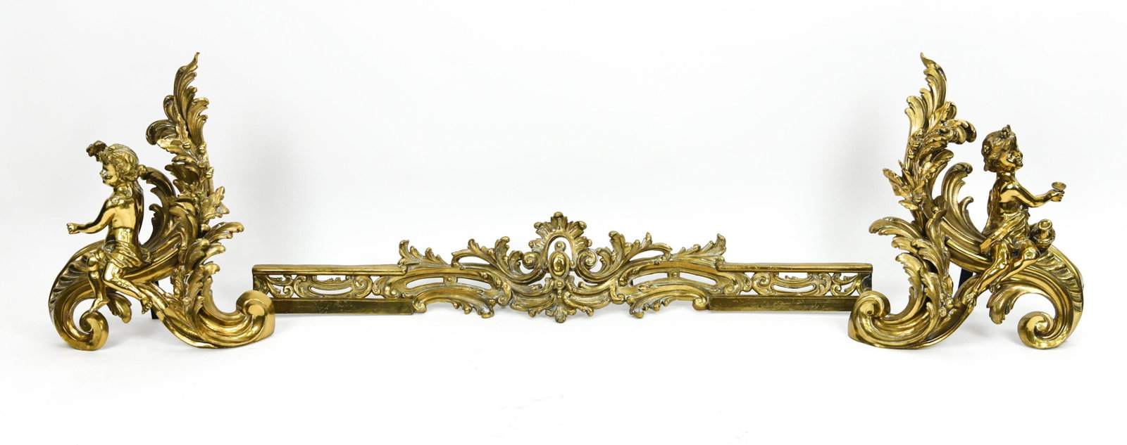 ORNATE GILT PUTTI FIREPLACE CHENET & FENDER: Highly ornate fireplace set consisting of a fender and a pair of putti fireplace chenet or andirons in gilt metal and brass. Fender stamped FC with indistinct character marks underneath; one andiron a